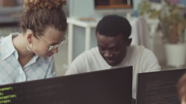 Multiethnic couple of software developers having conversation at workplace, sitting at office desk in front of computer monitors with program codes on black background