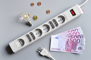 Euro banknotes, and cents, light bulb with power plug of an extension socket on gray surface. Concept for the rising cost of electricity. Expensive energy bill and rising electricity prices.