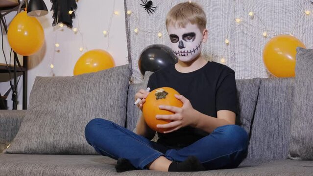 Child with makeup on his face paints a pumpkin for Halloween.