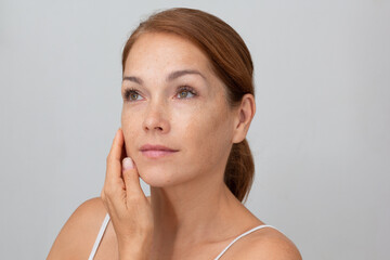 Portrait of cropped caucasian middle aged woman face with freckles holding fingers on cheek , showing soft skin on white background looking aside - obrazy, fototapety, plakaty