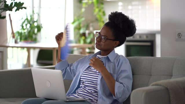 Exhausted African American woman suffering from summer weather using paper fan after heatstroke. Tired lady sits on sofa with laptop on lap in living room resting after physical activity causing sweat