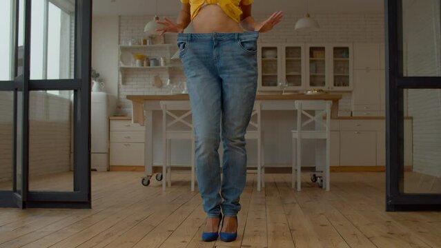 Low section of graceful elegant African American woman wearing stylish high heels and too large jeans, demonstrating success of weight loss diet program while standing in domestic room.