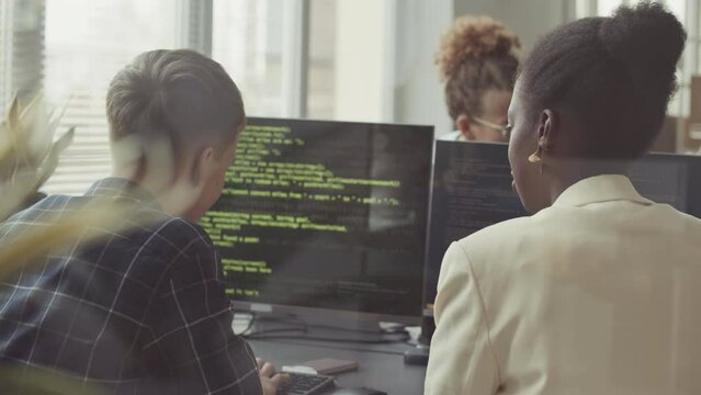 Rear view slowmo of diverse couple of programmers talking while writing program codes on PCs in office
