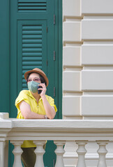 Adult female tourist in vintage style and protective face mask talking on smartphone while standing on terrace of retro building in vertical frame