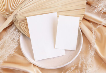 Blank cards on plate on dried palm leaf and beige silky fabric close up, greeting or wedding mockup