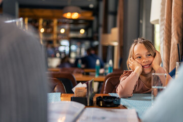 Portrait of beautiful smiling little girl sitting in restaurant with his parents waiting for launch.