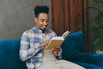 Young student fun man wear casual shirt sits on blue sofa couch read novel stories book stay at home hotel flat rest relax spend free spare time in living room indoors grey wall People lounge concept