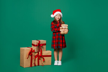 Full body merry little child kid girl 7 year old wears red dress Christmas hat posing near present box gift ribbon bow hold teddy bear isolated on plain green background Happy New Year 2023 concept