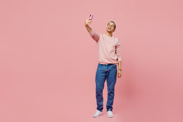 Full body young gay man wear sweatshirt hat do selfie shot on mobile cell phone post photo on social network isolated on plain pastel light pink color background studio. Lifestyle lgbtq pride concept.