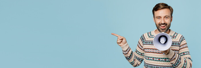 Young surprised man 30s wear sweater hold scream in megaphone announces discounts sale Hurry up point index finger aside isolated on plain pastel light blue cyan background People lifestyle concept.