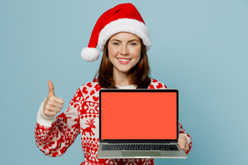 Young IT woman wear red sweater Santa hat posing work hold use laptop pc computer blank screen workspace area show thumb up isolated on plain blue cyan background. Happy New Year 2023 holiday concept.