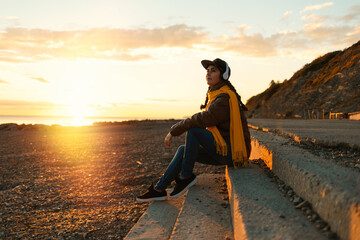 A young hipster Caucasian woman in a yellow scarf, cap and headphones, sitting on the steps and listening to music. Side view. Copy space. The concept of psychology and freedom