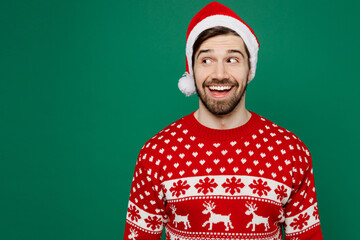 Merry smiling happy fun young man 20s wear red warm knitted sweater Santa hat posing look aside on workspace isolated on plain dark green background studio. New Year 2023 holiday celebration concept.