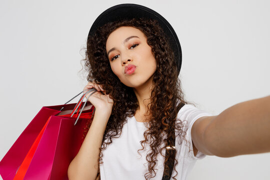 Close up young woman in t-shirt hat hold in hand red paper package bag after shopping do selfie shot pov mobile cell phone isolated on plain solid white background Black Friday sale buy day concept