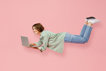 Full body side view young fun happy IT woman 20s she wear green shirt white t-shirt fly fall hover over air hurry up hold use work on laptop pc computer isolated on plain pastel light pink background.