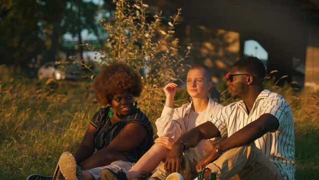 Three friends - two black people and one queer white person enjoying beautiful sunset during outdoor picnic. Diverse group of friends. Summer leisure time activities. High quality 4k footage