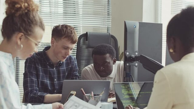Young diverse team of programmers writing program codes and chatting while coworking in modern office of successful IT company