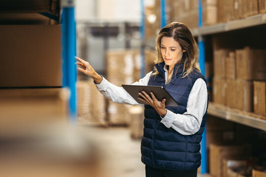 Female Manager Using A Digital Tablet While Doing A Stock Take In A Warehouse. Woman Checking SKU's Against An Inventory Checklist On A Warehouse Management System.