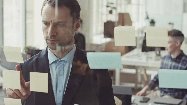 Serious Caucasian businessman in formalwear peeling sticky note with tasks to do off glass window while working in IT company