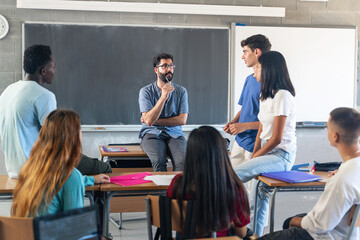 Group of teenagers with young male teacher at classroom, sitting talking and discussing together - obrazy, fototapety, plakaty
