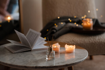 Home liquid fragrance with scented candle and open paper book over christmas glow lights and chair...