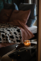 Scented burning candle stay on wooden tray and knitted textile in dark room over bed. Cozy home atmosphere lifestyle.