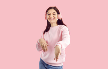 Happy carefree young woman in massive wireless headphones having fun dancing on pink background....