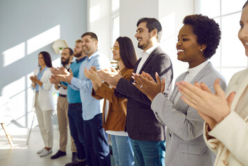 Happy office team demonstrates pleasure of business training by joyfully applauding speaker. Side view of satisfied multiracial colleagues standing in row in bright office and clapping their hands. - Powered by Adobe