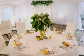 Festive table with a white tablecloth, and dishes in tent. The Banquet hall decorated flowers, leave, herbs in vase, citrus fruits lemons. Chairs and tables with atmospheric decor in the backyard.