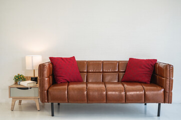 Brown leather sofa with two red pillows in front of white wall in living room with side table and lamp.