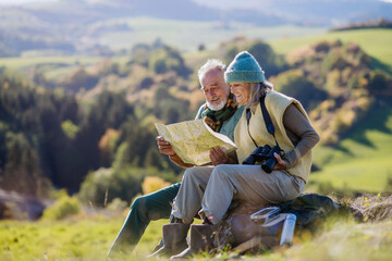 Senior couple having break, looking into paper map during hiking in autumn nature.
