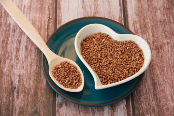 Small grains of natural buckwheat in a decorative plate in the form of a heart and in a wooden spoon