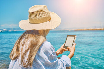 Fototapeta premium Fashion pretty woman outdoors lifestyle watching, reading on tablet ebook on the beach in summer day. Wearing wide brimmed hat, Sunbating with uv protection. Concept of beach vacation.