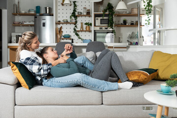 Happy married homosexual female gay couple laughing and embracing on the sofa with smile on their faces. Lesbian couple at home enjoying life together in their new apartment.