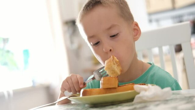 Funny little Caucasian boy eats croutons with a fork while sitting at table at home in kitchen. Delicious nutritious breakfast for a child.
