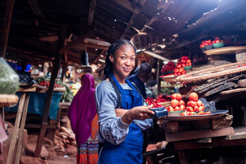 Market woman using a pos terminal for buying and selling in aa typical local african tomatoes and vegetables market - obrazy, fototapety, plakaty