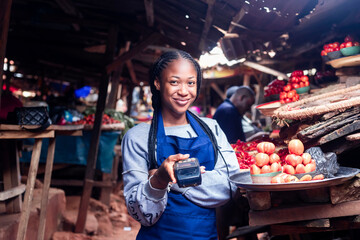 African millennial lady wearing colourful Apron selling tomatoes and vegetables in a typical local african market holding a POS terminal - obrazy, fototapety, plakaty
