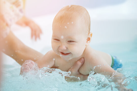 Father holds excited baby daughter tummy learning to swim in bath water. Baby girl looks with smile enjoying washing in bathtub with father help, sunlight