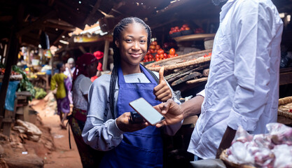 A young African friendly saleswoman passing pos terminal over counter to customer paying with smartphone Smiling given a thumbs up - obrazy, fototapety, plakaty