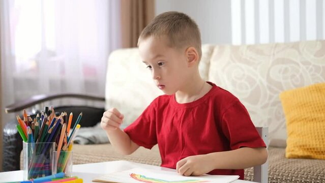 Portrait of funny happy little caucasian boy sitting at table and drawing with colored pencils and markers. Art therapy, rehabilitation, leisure.