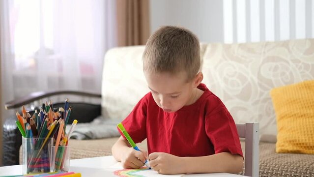 Portrait of serious little caucasian boy with autism sitting at table and drawing with colored pencils. Art therapy, rehabilitation, leisure.