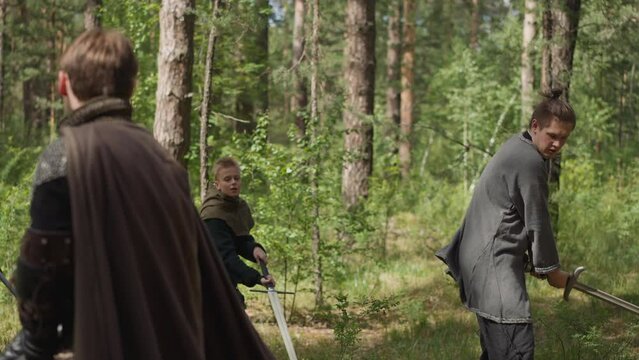 Teenage boys train sword strikes with master in sunny pine forest at quest slow motion. Historical reconstruction club for children. Role playing