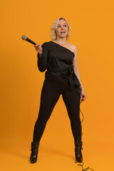 blonde woman in a black blouse with a bare shoulder with a wired microphone in her hand on a yellow background