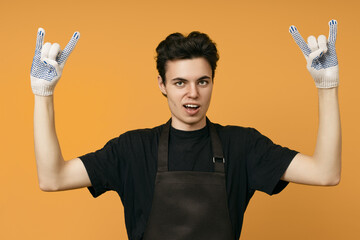 young man in a black t-shirt and apron in work gloves shows a rocker goat sign with his hands