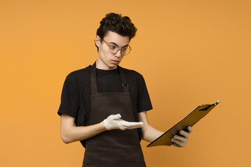 a young man in a black t-shirt and apron in work gloves is perplexed and looks at a tablet for papers in his hands