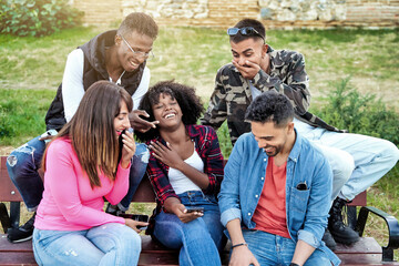 Multi-ethnic friends having fun and laughing while using a mobile phone sitting on a bench outdoors. Technology and friendship concept.