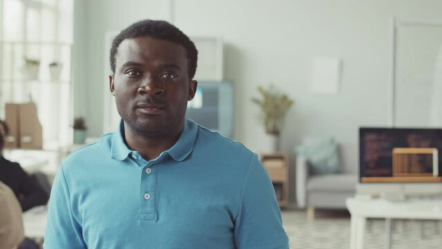 Chest up slowmo portrait of young Black man in blue polo shirt looking at camera standing at modern programmer office