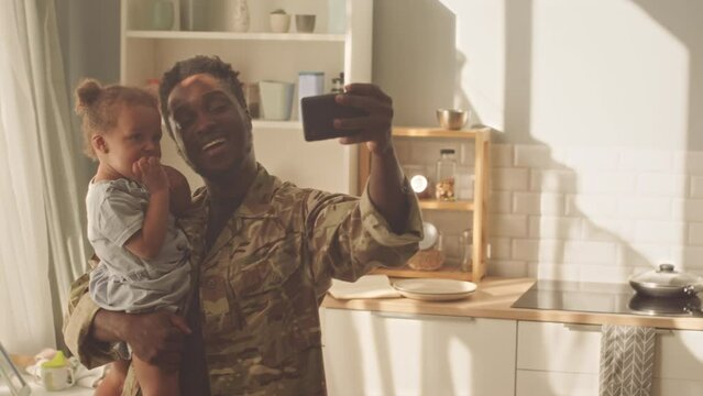 Cheerful African American man taking selfie portrait on smartphone with his pretty toddler girl holding her in arms at kitchen