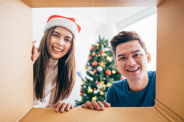 Young Asian couple opening a Christmas present together with a smile, views from inside of the box with a decorated Christmas tree in the background. Boxing day during the Christmas holiday.