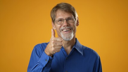 Portrait of mature man in blue shirt having idea moment giving thumbs up on solid yellow studio background. Smiling happy guy showing eureka